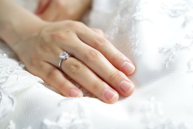 Close-up of a bride's hand resting on a wedding dress, showcasing a beautifully delicate engagement ring with a diamond centerpiece. This can be used for wedding blogs, bridal magazines, jewelry advertisements, and romantic themed content.