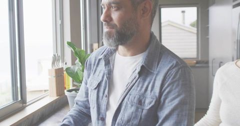 Bearded man gazing out kitchen window with partner at sunlit countertop, minimalist home