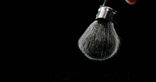 A makeup brush is being held by a person against a black background, with powder particles visibly scattering from its bristles, with copy space. This action captures the dynamic movement of makeup application or the cleaning of the brush.