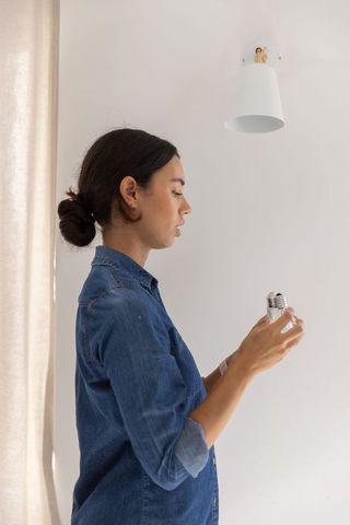 Woman installing led bulb into wall lamp near curtain minimalist home renovation
