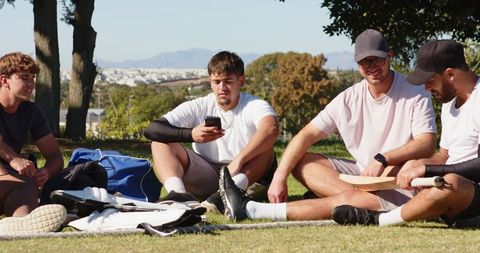 Young men relaxing after cricket match, sitting on grass with bat, pads and smartphone
