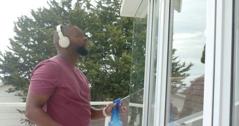 African american man wearing headphones cleaning glass sliding door on coastal balcony