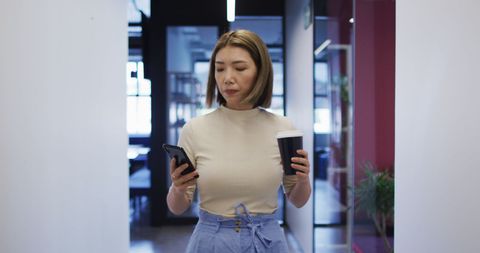Businesswoman walking in office holding coffee and smartphone