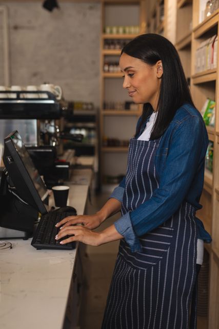 Smiling biracial female cafe owner using desktop computer at counter in a coffee shop. She is wearing an apron and appears to be managing her business operations. This image can be used for themes related to small business, entrepreneurship, customer service, and technology in the food and beverage industry.