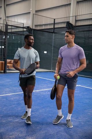 Male friends walking and talking on indoor padel court holding paddles and ball