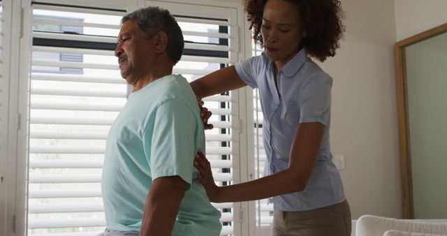Medical professional assisting senior man at home with back pain. Useful for articles on elderly care, home health care, therapy, physical rehabilitation, and senior patient support.