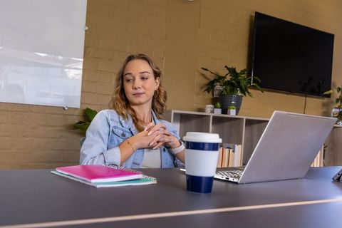 Contemplative young woman sitting at desk with laptop and coffee in modern office space