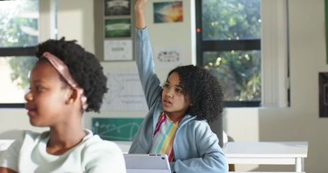 African American teen student raising hand while using tablet in bright classroom