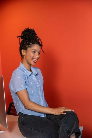 Smiling african american woman sitting on desk in orange workspace wearing polka-dot blouse