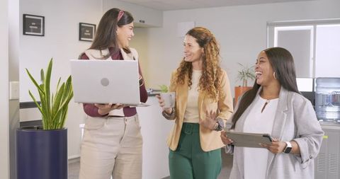 Diverse women walking and talking in modern office holding laptop, tablet and coffee cup