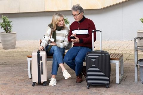 Senior couple checking phone while waiting with luggage and coffee on urban bench