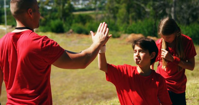 Coach high-fiving young boy during outdoor sports event on sunny day. Boy and girl both dressed in red shirts with green background. Scene depicts sportsmanship and teamwork, perfect for use in articles about physical education, youth sports, and coaching techniques.