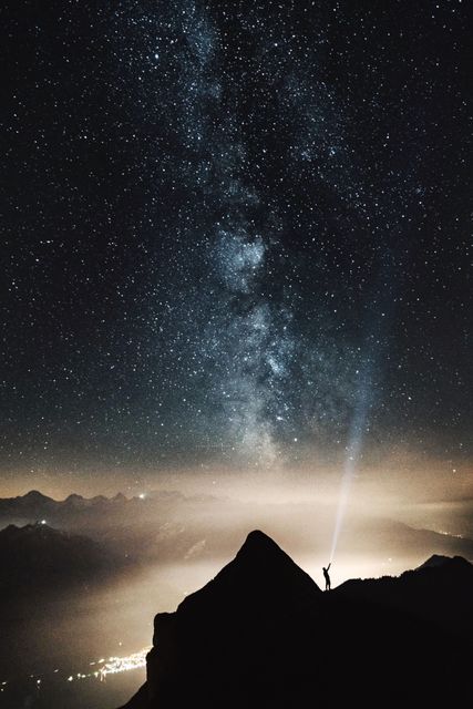 This image shows an astronomer standing on a mountain peak, pointing a flashlight up towards the starry night sky, with the Milky Way clearly visible. The scene is illuminated softly by the distant city lights and ambient atmosphere, creating a serene and awe-inspiring view. Suitable for use in themes related to astronomy, nature, adventure, and outdoor exploration. Ideal for travel publications, educational materials, motivational posters, or backgrounds for nature-related subjects.