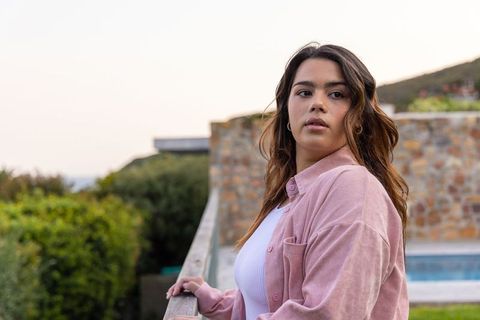 Contemplative young woman leaning on terrace railing overlooking pool and garden at sunset