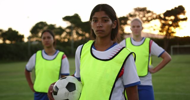 Three female soccer players training on a field at sunset, wearing neon yellow practice pinnies. Ideal for use in content related to sports training, teamwork, women's athletics, youth programs, and inspiration in sports.