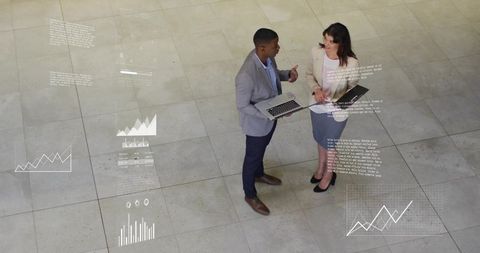 Black man and white woman discussing data with laptop and projected charts in atrium