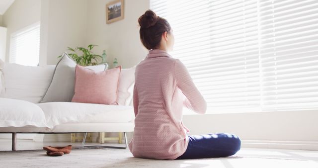 Woman sitting on floor in living room, meditating and basking in sunlight streaming through window blinds. Ideal for use in articles about meditation, mindfulness, home relaxation techniques, and stress relief. Conveys tranquility and peaceful mental state.