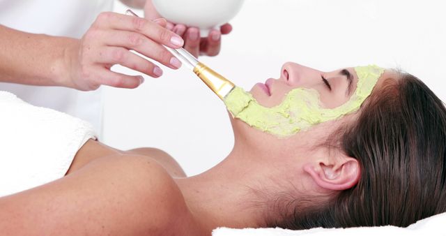 A young Caucasian woman is receiving a facial treatment with a green mask being applied by a beautician, with copy space. Spa and wellness concepts are illustrated by the skincare procedure being performed.