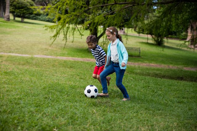 Two children, a boy and a girl, are playing soccer on a grassy field in a park. They are both focused on the ball, enjoying an outdoor activity. This image can be used for promoting outdoor activities, sports for kids, family fun, and healthy lifestyles.