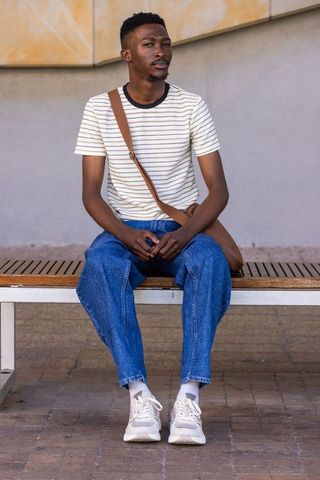 Stylish african man sitting on bench wearing striped tee, denim and crossbody bag
