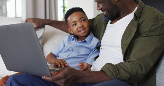 This image depicts a father and son sitting together on a sofa and using a laptop. They are smiling, suggesting they are enjoying their time together. The warm and inviting indoor environment makes it a perfect shot for themes focusing on family bonding, parenting, and home education. Ideal for use in articles, advertisements, and blog posts related to family relationships, technology in everyday life, and promoting positive father-son activities.