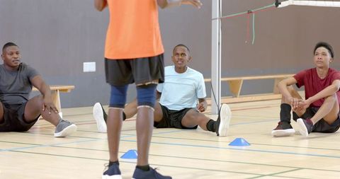 Diverse male athletes sitting on indoor court listening to coach during team training drill