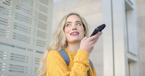 Young woman speaking into smartphone while commuting in mustard sweater near modern facade