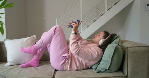 Young woman reclining on sofa in pink loungewear listening to music on smartphone