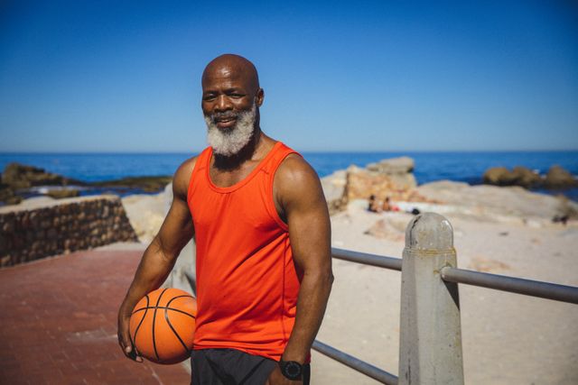 Senior African American man holding a basketball while standing on a promenade by the beach. He is wearing a bright orange tank top and smiling, showcasing an active and healthy lifestyle. This image can be used for promoting senior fitness, retirement activities, healthy living, and outdoor sports.