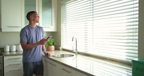 African american man smiling while holding smartphone at sunlit kitchen window