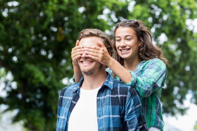 Young woman playfully covering man's eyes while standing outdoors in a lush green environment. Both are smiling and dressed in casual plaid shirts. Ideal for use in advertisements, relationship blogs, social media posts, and lifestyle articles focusing on love, joy, and togetherness.