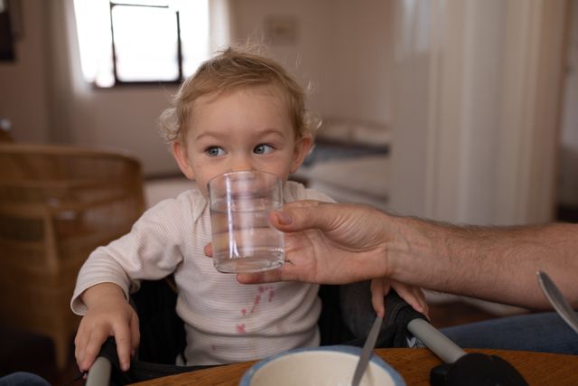 Front view of a Caucasian baby sitting on a high chair by the table in the dining room, wearing white shirt, drinking water held by his father.