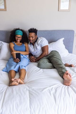 Relaxing together: diverse black couple lounging on bed, smiling and scrolling smartphone