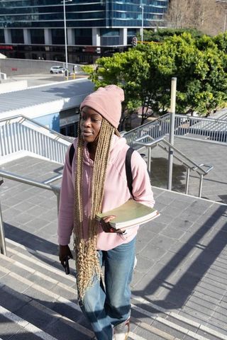 Female student walking up city stairs wearing pink beanie and hoodie carrying notebooks