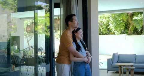 Couple embracing at sliding glass door overlooking sunny backyard pool and modern patio