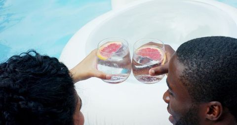 Poolside toasting: couple relaxing on white float raising grapefruit cocktail glasses
