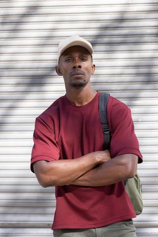 African american man standing with arms crossed wearing maroon tee and beige cap