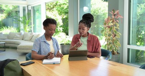 African american mentor tutoring teen girl using tablet and notebook in bright home study