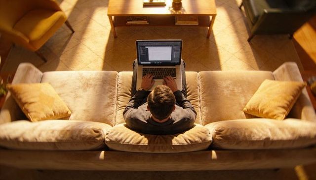 Overhead view of man typing on laptop on sunlit beige sofa in cozy living room workspace