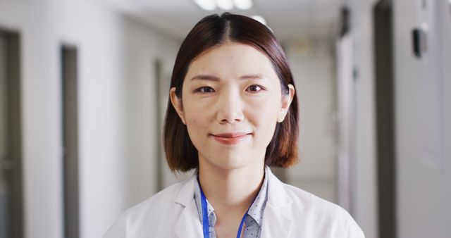 Female Asian doctor stands in hospital corridor, confidently smiling. Ideal for promoting healthcare services, medical websites, hospital brochures, professional profiles of medical staff, or health-related articles.