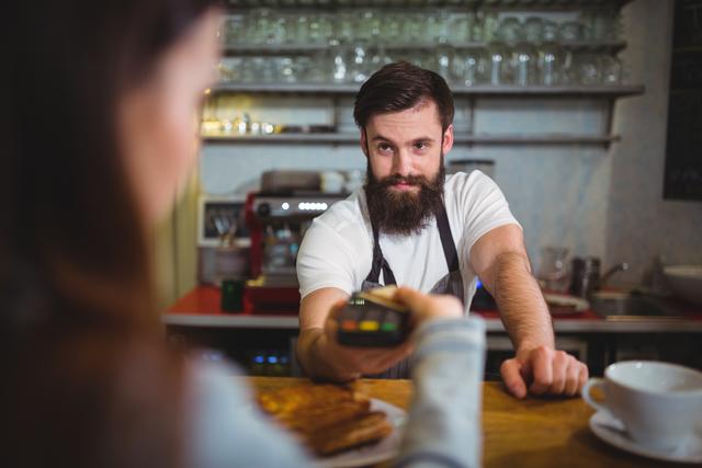 Bearded barista holding payment terminal while a customer makes a payment at a cafe counter. Ideal for illustrating concepts related to retail transactions, electronic payments, customer service, small business operations, and food and beverage industry. Great for use in marketing materials, blogs about modern payment solutions, or advertisements for cafes and coffee shops.