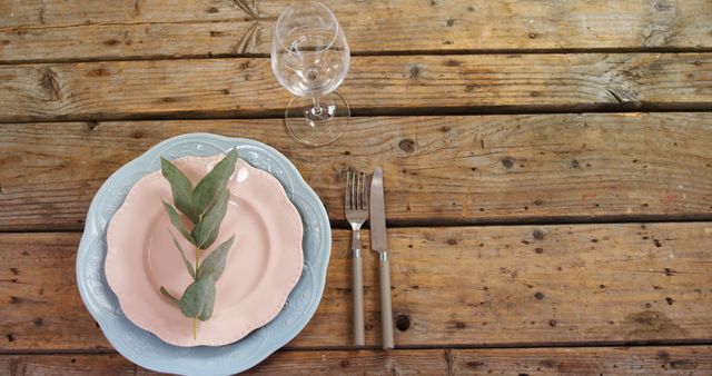 A rustic table setting features a pink napkin, silverware, and an empty wine glass on a wooden table, with copy space. The arrangement suggests a romantic or celebratory outdoor dining experience.