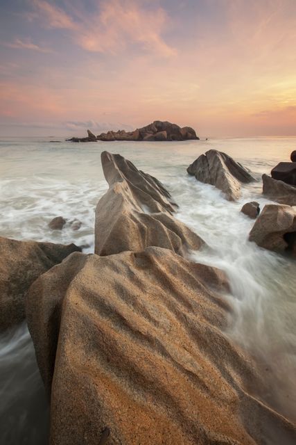 Depicting a serene coastal landscape during sunrise with detailed rock formations in foreground and the pastel-colored sky meeting the calm ocean. Useful for travel brochures, website backgrounds, inspirational content, and nature-related publications conveying tranquility and natural beauty.