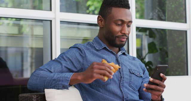 Man wearing denim shirt sitting outdoors, enjoying sandwich while using smartphone. Ideal for content on modern lifestyle, technology impact, outdoor dining, and multitasking.