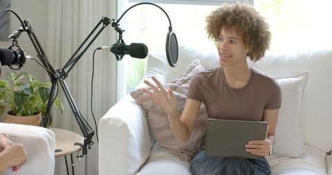 African american woman hosting cozy home podcast on sofa with microphones and tablet