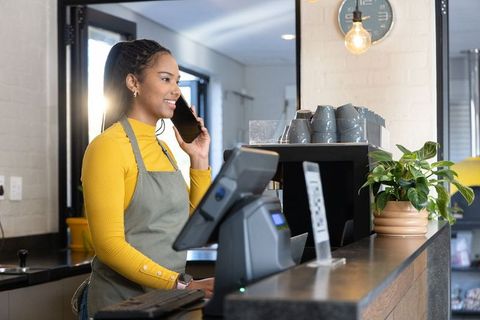 Smiling female barista talking on smartphone behind modern coffee shop counter with pos
