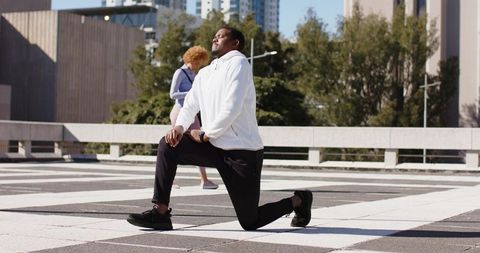 African American man performing kneeling lunge on rooftop plaza, woman walking nearby