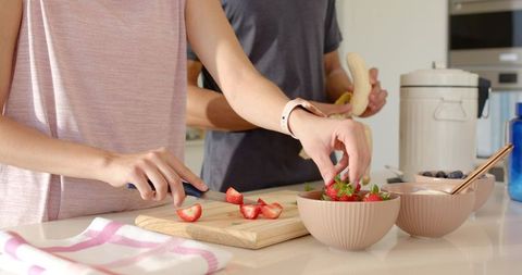Couple preparing fresh fruit on kitchen counter, slicing strawberries and peeling banana