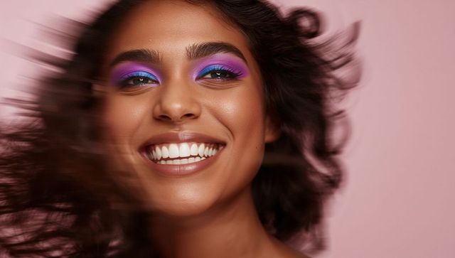 Vibrant purple and blue eyeshadow on smiling woman with windblown hair, glossy lips