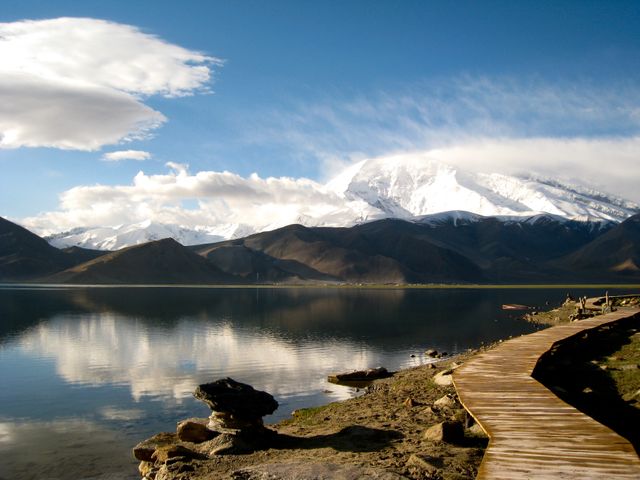Breathtaking view of a tranquil lake surrounded by snow-capped mountains with a wooden path leading toward the water. Reflection on the calm water adds to the serenity of the scene. Ideal for use in travel magazines, nature blogs, desktop wallpapers, and promotional materials for outdoor adventures.